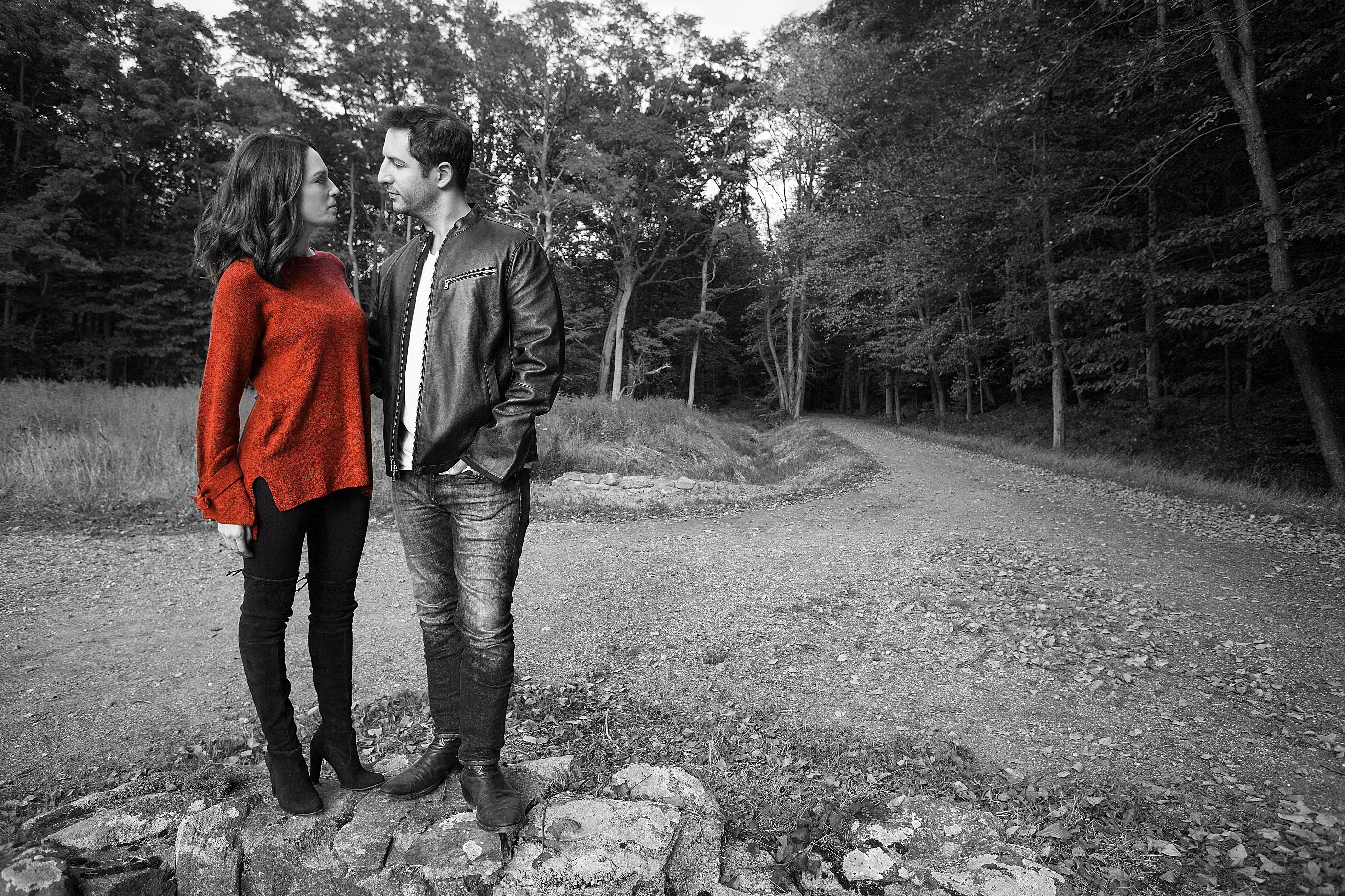 Engagement photo of couple standing on forest path crossroads, woman in red sweater gazing at partner